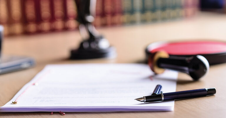 Auf einem Tisch liegen ein Kaufvertrag, ein Füller und ein Stempel, im Hintergrund stehen Rechtsbücher | Notartermin
