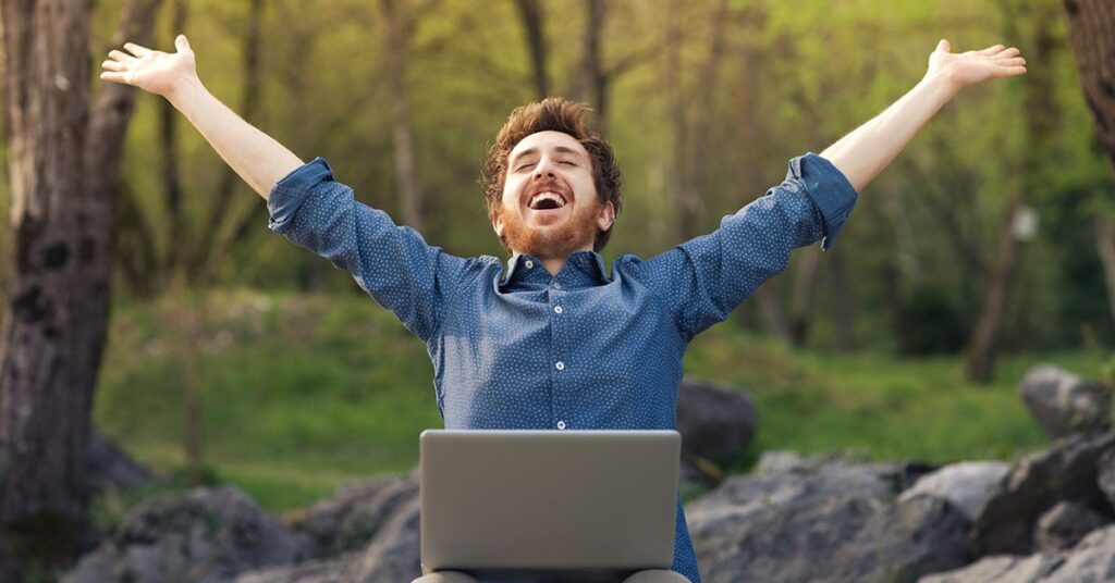 Ein Mann mit Laptop auf - Beruflicher Wechsel dem Schoß im Wald wirft glücklich die Arme hoch und freut sich über neuen Job