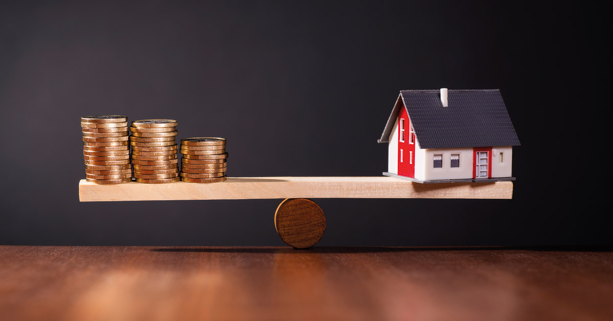 Auf einem Holztisch steht eine Waage aus Holz, die ein Haus gegen Geld abwiegt vor einem dunklen Hintergrund | Immobilienkauf Inflation Zinsen
