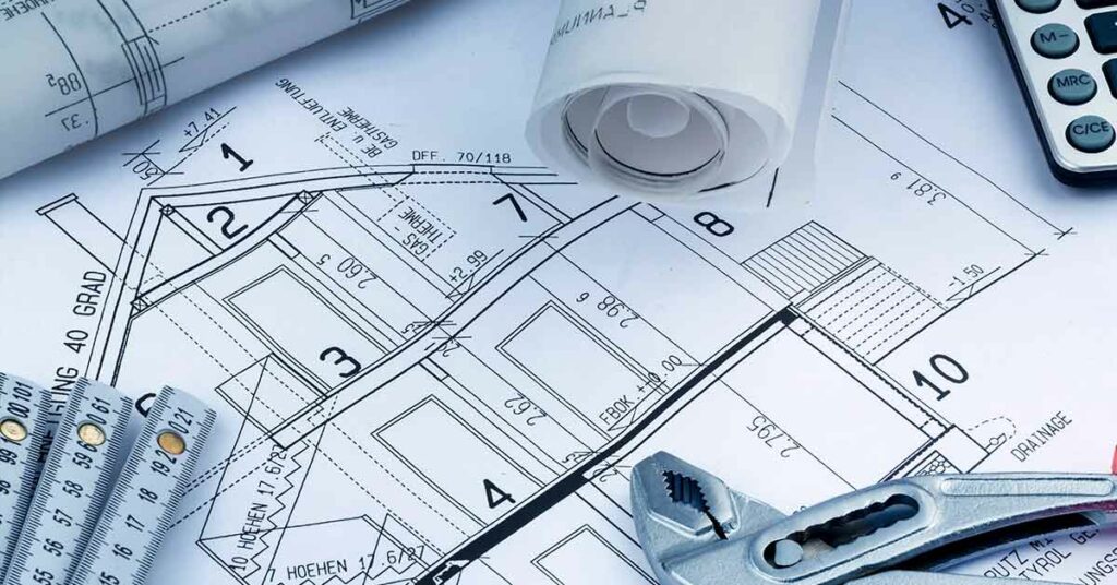Grundrissplan eines Hauses, darauf liegen ein Zollstock, eine Rohrzange, ein Taschenrechner und gerollte Baupläne | Barrierefreier Umbau