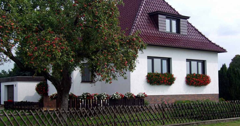 Ein Haus mit weißer Fassade und rotem Dach, mit einem Baum im Vorgarten und einem Holzzaun zur Straße | Fehler Immobilienverkauf