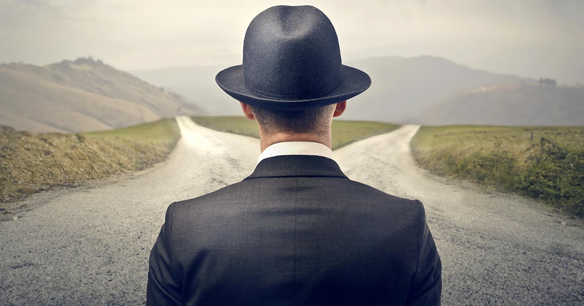 Ein Mann mit Anzug und Hut, steht an einer Weggabelung, vor der Entscheidung, ob er eine Immobilie kaufen oder mieten soll | Mieten oder kaufen