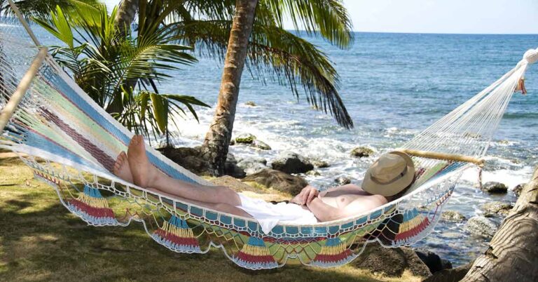 Ein Mann liegt entspannt in seiner Hängematte am Strand im Ausland unter Palmen, mit Hut auf dem Gesicht, und genießt sein Leben, weil er weiß, dass ein Qualitätsmakler sich um den Verkauf seiner Immobilie in Deutschland kümmert | Immobilienverkauf