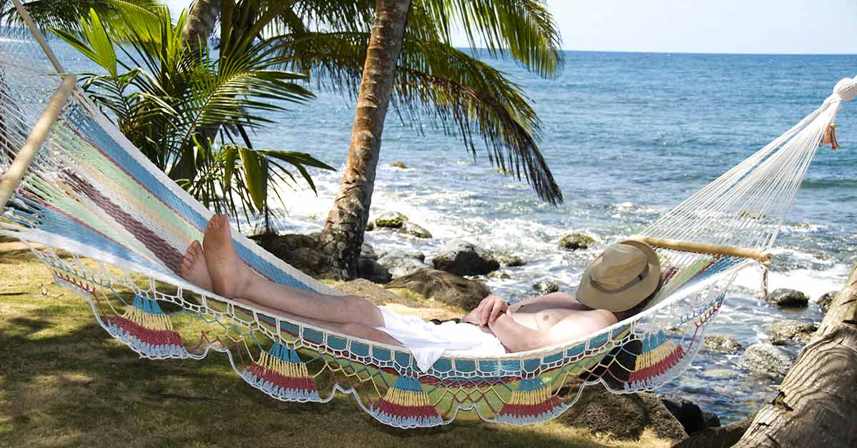 Ein Mann liegt entspannt in seiner Hängematte am Strand im Ausland unter Palmen, mit Hut auf dem Gesicht, und genießt sein Leben, weil er weiß, dass ein Qualitätsmakler sich um den Verkauf seiner Immobilie in Deutschland kümmert | Immobilienverkauf