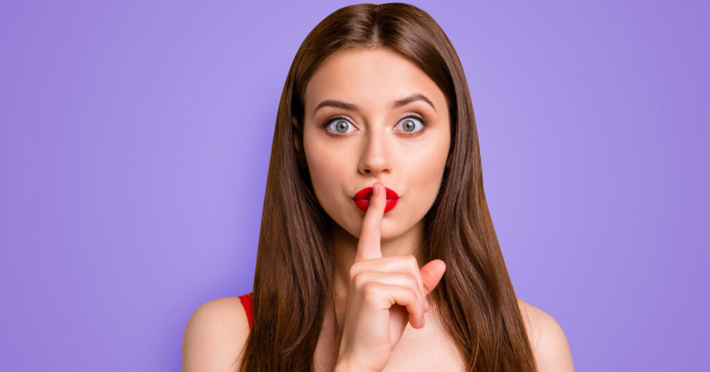 ein junge Frau, die sich den Zeigefinger vor den Mund hält um Verschwiegenheit zu demonstrieren