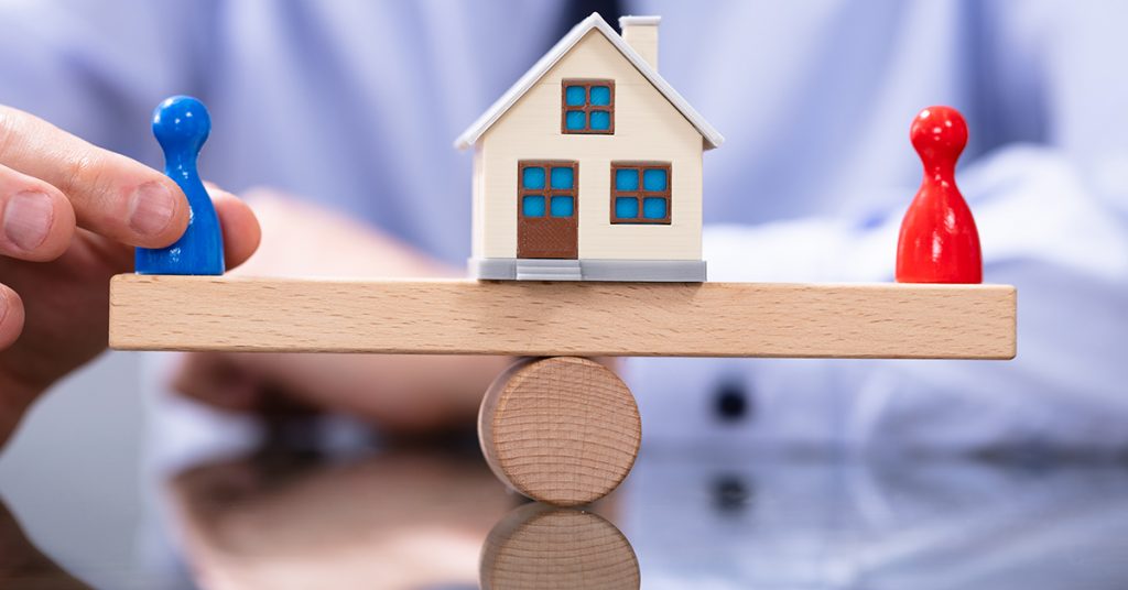 Ein Häuschen auf der Waage mit zwei Figuren in rot und blau - Scheidungsimmobilie